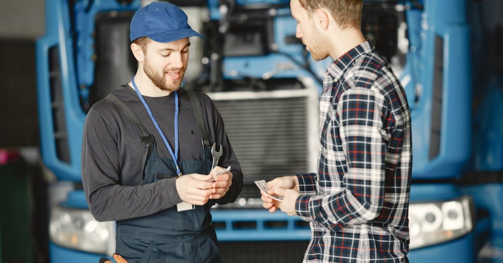 Auto mechanic in blue uniform receives payment from customer in a garage setting with a truck in the background.